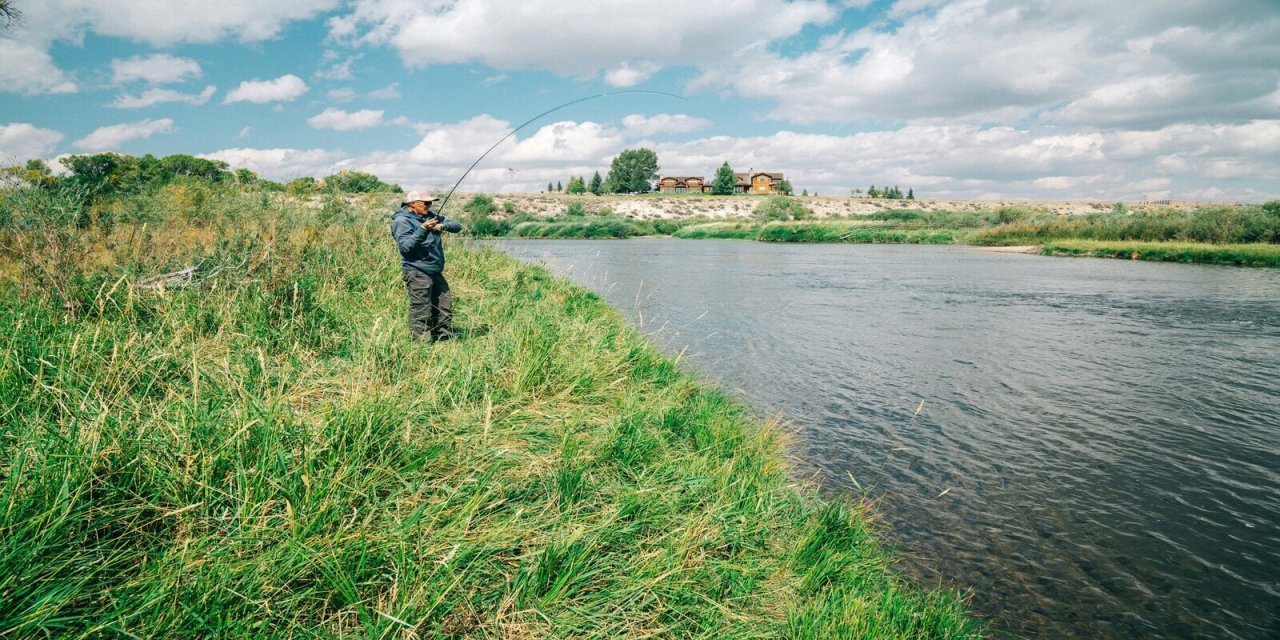 Large field by the water with a man in a blue jacket fishing. Blue sky with white clouds. 
