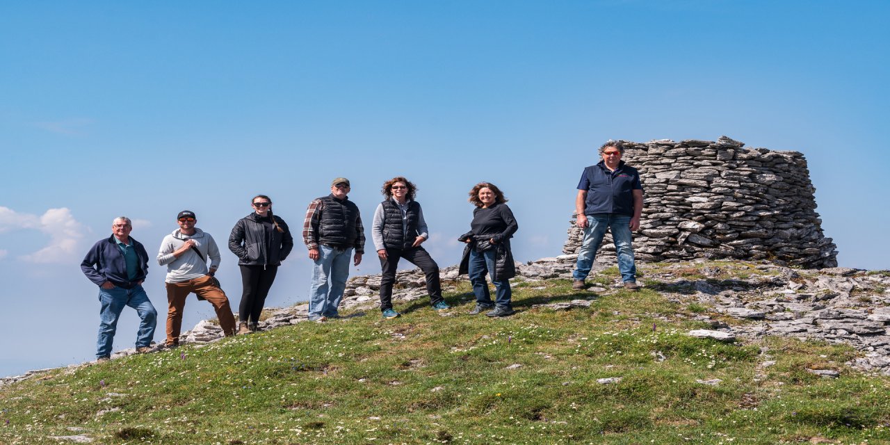 Tour group and guide standing on a green mountain side posing beside a small stone tower. The sky is blue and the grass is earthy green